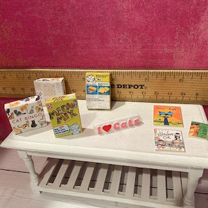 K&ouml;nnte beinhalten: Eine Sammlung von Miniaturartikeln zum Thema Katzen, darunter eine Cat Bingo-Schachtel, eine Meow Mix-Schachtel, eine gelbe Schachtel, ein wei&szlig;es I Love Cats-Schild und drei kleine B&uuml;cher mit den Titeln Pete the Cat, The Bookshop Cat und ein Katzenportr&auml;t.
