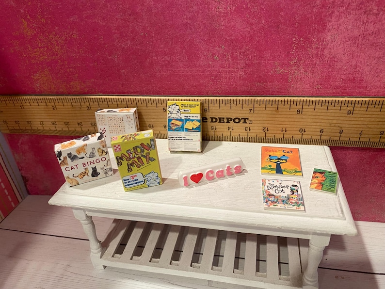 K&ouml;nnte beinhalten: Eine Sammlung von Miniaturartikeln zum Thema Katzen, darunter eine Cat Bingo-Schachtel, eine Meow Mix-Schachtel, eine gelbe Schachtel, ein wei&szlig;es I Love Cats-Schild und drei kleine B&uuml;cher mit den Titeln Pete the Cat, The Bookshop Cat und ein Katzenportr&auml;t.