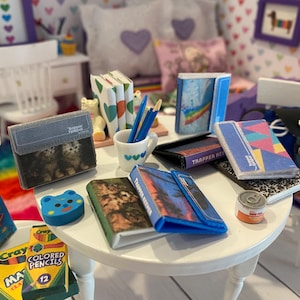 May include: A miniature dollhouse scene featuring a white table with school supplies. There are small books, notebooks, pencils, and a pencil case with a kitten image. A blue eraser and a can of food are also on the table.