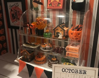 May include: A miniature Halloween-themed display with a glass case filled with various treats, including cakes, candy, and cookies. The case is decorated with a banner that reads "Halloween". There is a calendar with the month of October highlighted, and a small pumpkin figurine.