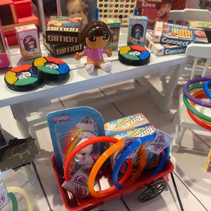 Op de afbeelding: Een miniatuursc&egrave;ne met diverse speelgoed en spelletjes. Bevat een Barbie-pop, een Dora the Explorer-figuur en verschillende bordspellen zoals Simon en Connect Four. Er zijn ook kleurrijke ringen, een rode wagen en een Lite-Brite-speelgoed.
