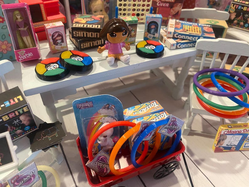 Op de afbeelding: Een miniatuursc&egrave;ne met diverse speelgoed en spelletjes. Bevat een Barbie-pop, een Dora the Explorer-figuur en verschillende bordspellen zoals Simon en Connect Four. Er zijn ook kleurrijke ringen, een rode wagen en een Lite-Brite-speelgoed.