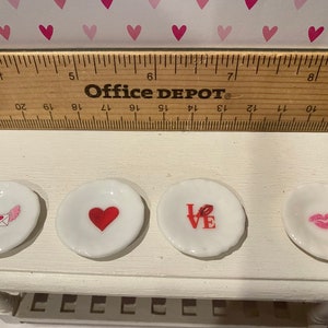 May include: Four small white ceramic plates with Valentine-themed graphics including a winged envelope, a red heart, the word LOVE with a lip print, and a pair of pink lips. A wooden ruler is positioned above the plates for scale reference.