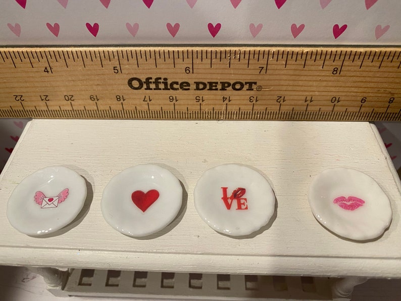 May include: Four small white ceramic plates with Valentine-themed graphics including a winged envelope, a red heart, the word LOVE with a lip print, and a pair of pink lips. A wooden ruler is positioned above the plates for scale reference.