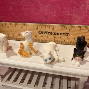 K&ouml;nnte beinhalten: Vier Miniatur-Katzenfiguren auf einem wei&szlig;en Tisch vor einem Office Depot-Lineal. Die Katzen umfassen eine rein wei&szlig;e Katze, eine orange-wei&szlig; getigerte Katze, eine wei&szlig;e Katze mit blauen Augen und eine schwarz-wei&szlig;e Katze mit gelben Augen.