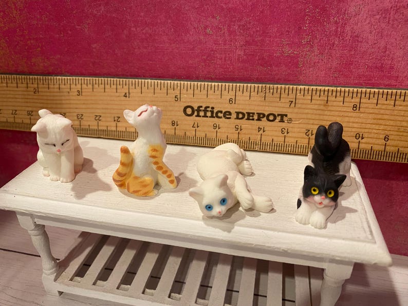 K&ouml;nnte beinhalten: Vier Miniatur-Katzenfiguren auf einem wei&szlig;en Tisch vor einem Office Depot-Lineal. Die Katzen umfassen eine rein wei&szlig;e Katze, eine orange-wei&szlig; getigerte Katze, eine wei&szlig;e Katze mit blauen Augen und eine schwarz-wei&szlig;e Katze mit gelben Augen.
