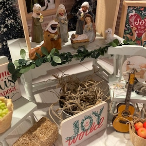 May include: A miniature Christmas nativity scene with figurines of Mary, Joseph, and baby Jesus. The scene is set on a white table with a green garland and a sign that reads "Joy to the World".