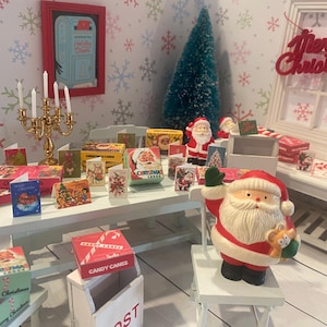 May include: A miniature Christmas scene with a white table displaying holiday cards, candy boxes, and Santa Claus figurines. A red "Merry Christmas" sign hangs in the background, with a small Christmas tree and a candelabra.