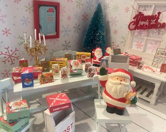 May include: A miniature Christmas scene with a white table displaying holiday cards, candy boxes, and Santa Claus figurines. A red "Merry Christmas" sign hangs in the background, with a small Christmas tree and a candelabra.