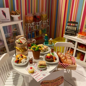 May include: A miniature dollhouse kitchen set with a white table and chairs, a variety of miniature food items, including sandwiches, pastries, and fruit, and a cash register. The kitchen is decorated with colourful striped wallpaper and a sign that reads "Canary Cafe".