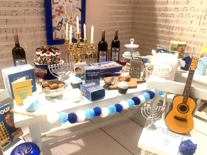 May include: A white table set for a Hanukkah celebration with a menorah, candles, wine bottles, a guitar, and a blue and white garland. The table is decorated with a blue and white garland, a menorah, candles, wine bottles, a guitar, and a cake. The table is set for a Hanukkah celebration.