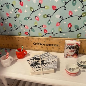 May include: A miniature Christmas scene with a white table, two miniature mugs, a box of Corn Flakes cereal, a newspaper clipping, and two small bowls with spoons. The background is a blue and white patterned paper with red and white lights.