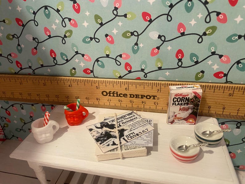 May include: A miniature Christmas scene with a white table, two miniature mugs, a box of Corn Flakes cereal, a newspaper clipping, and two small bowls with spoons. The background is a blue and white patterned paper with red and white lights.