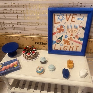 May include: A white table with a blue and white menorah, a blue dreidel, a white dreidel, a gold dreidel, a blue cake stand, a chocolate cake with cherries, a box of jelly donuts, a blue cupcake, a blue donut, and a blue and white cupcake. The table is in front of a blue picture frame with the words "Love and Light" and a Star of David.
