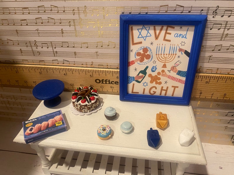 May include: A white table with a blue and white menorah, a blue dreidel, a white dreidel, a gold dreidel, a blue cake stand, a chocolate cake with cherries, a box of jelly donuts, a blue cupcake, a blue donut, and a blue and white cupcake. The table is in front of a blue picture frame with the words "Love and Light" and a Star of David.