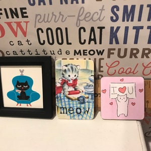 May include: Three framed pictures with cat-themed designs. The first picture features a black cat sitting in a blue armchair with a gray mouse on top. The second picture shows a cartoon cat baking in a kitchen. The third picture is pink with a white cat holding a sign that says "I love you" with a red heart.