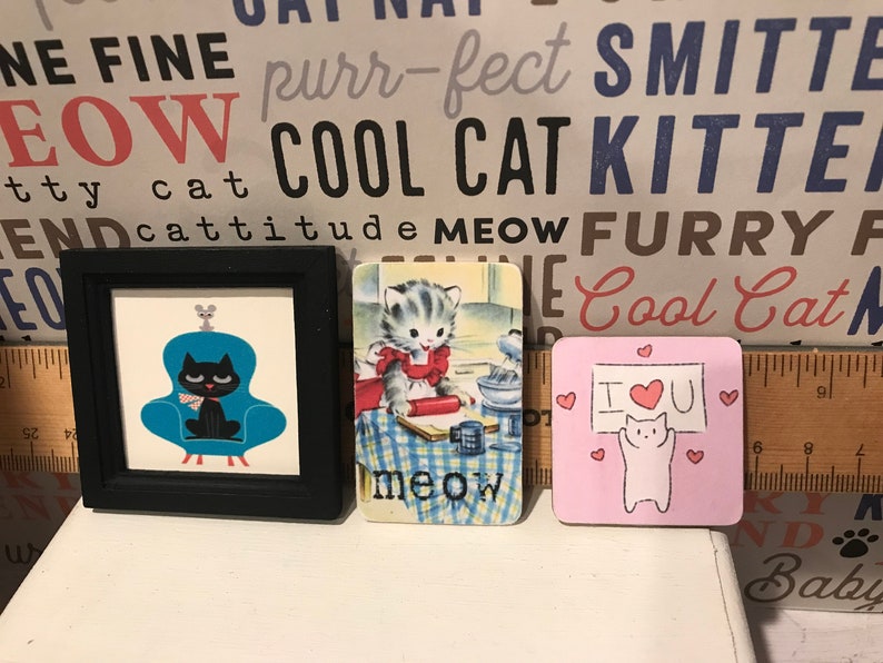 May include: Three framed pictures with cat-themed designs. The first picture features a black cat sitting in a blue armchair with a gray mouse on top. The second picture shows a cartoon cat baking in a kitchen. The third picture is pink with a white cat holding a sign that says "I love you" with a red heart.
