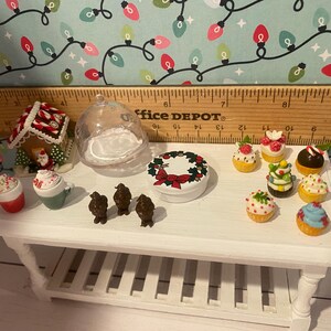 May include: A white table with a variety of miniature Christmas-themed decorations, including a gingerbread house, a wreath, a glass dome, and several cupcakes. The table is made of wood and has a white finish. The decorations are all in a festive Christmas style.