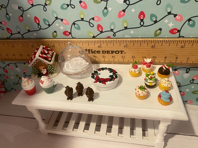 May include: A white table with a variety of miniature Christmas-themed decorations, including a gingerbread house, a wreath, a glass dome, and several cupcakes. The table is made of wood and has a white finish. The decorations are all in a festive Christmas style.