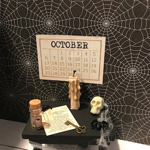 Pode incluir: Uma cena de Halloween em miniatura com uma mesa preta, uma caveira branca, uma aranha preta, uma vela, uma garrafa com uma rolha, uma chave e um pedaço de papel com o texto "A Witches Lucky Spell". A mesa está colocada sobre um fundo de teia de aranha preta e branca com um calendário para outubro.