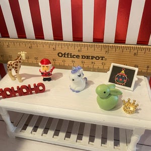 May include: A collection of miniature figurines on a white table, including a giraffe, Santa Claus, a unicorn, a green bunny, a small framed picture, and a gold crown. The word "HOHOHO" is also present. A wooden ruler is in the background.