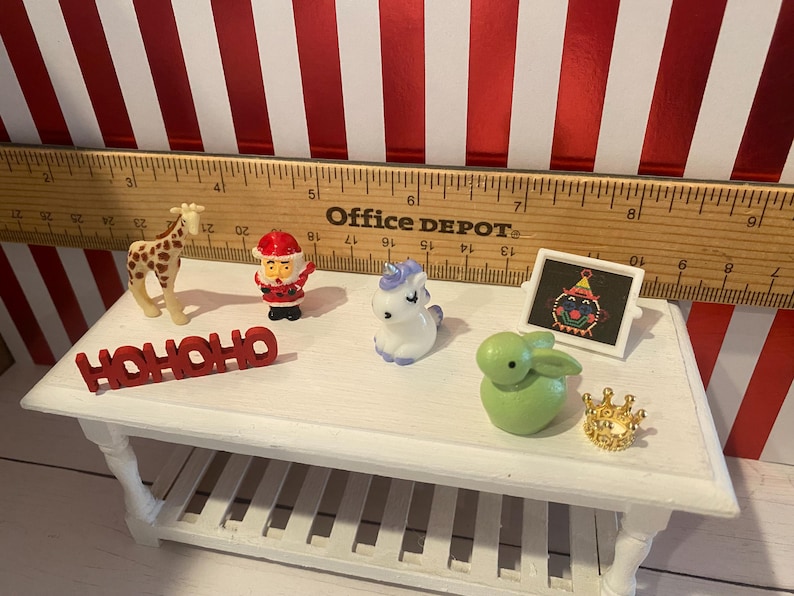 May include: A collection of miniature figurines on a white table, including a giraffe, Santa Claus, a unicorn, a green bunny, a small framed picture, and a gold crown. The word "HOHOHO" is also present. A wooden ruler is in the background.