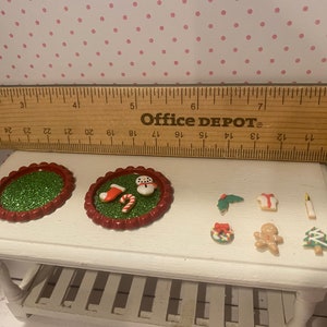 May include: A set of miniature Christmas decorations, including a snowman, a stocking, a candy cane, a gingerbread man, a wreath, a Christmas tree, and a candle. The decorations are on a white table with a green and red glitter base.