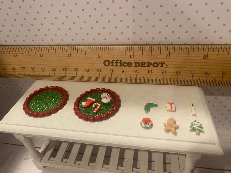 May include: A set of miniature Christmas decorations, including a snowman, a stocking, a candy cane, a gingerbread man, a wreath, a Christmas tree, and a candle. The decorations are on a white table with a green and red glitter base.