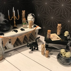 Puede incluir: Una escena de Halloween en miniatura con un gato negro, un caldero, velas y una calavera. La escena est&aacute; ambientada en una mesa blanca con una pancarta de arpillera y un fondo de telara&ntilde;a negra.