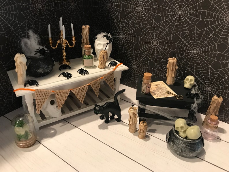 Puede incluir: Una escena de Halloween en miniatura con un gato negro, un caldero, velas y una calavera. La escena est&aacute; ambientada en una mesa blanca con una pancarta de arpillera y un fondo de telara&ntilde;a negra.