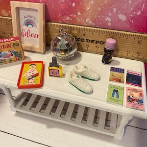 K&ouml;nnte beinhalten: Ein wei&szlig;es Regal mit einer Vielzahl von Miniaturartikeln, darunter ein Spiel namens Operation, ein kleines gerahmtes Schild mit der Aufschrift "Always Believe", eine Discokugel, eine kleine Flasche Fl&uuml;ssigkeit, ein Paar wei&szlig;e Turnschuhe und mehrere kleine B&uuml;cher mit bunten Umschl&auml;gen.