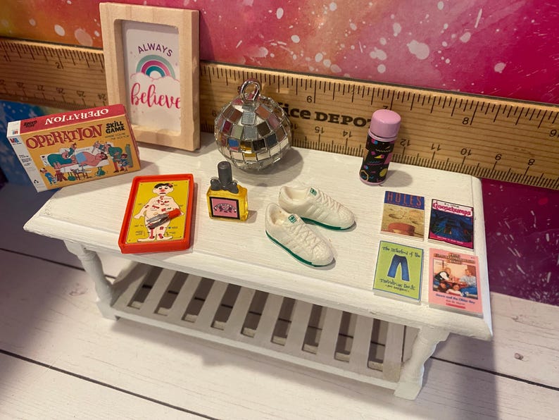 K&ouml;nnte beinhalten: Ein wei&szlig;es Regal mit einer Vielzahl von Miniaturartikeln, darunter ein Spiel namens Operation, ein kleines gerahmtes Schild mit der Aufschrift "Always Believe", eine Discokugel, eine kleine Flasche Fl&uuml;ssigkeit, ein Paar wei&szlig;e Turnschuhe und mehrere kleine B&uuml;cher mit bunten Umschl&auml;gen.