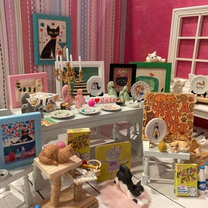 K&ouml;nnte beinhalten: Ein Miniatur-Puppenhauszimmer mit katzenthematischer Dekoration, darunter gerahmte Katzenbilder, kleine Katzenfiguren in verschiedenen Posen, winzige Teller, ein Miniatur-Kratzbaum und Schachteln mit der Aufschrift Meow Mix auf wei&szlig;en M&ouml;beln.