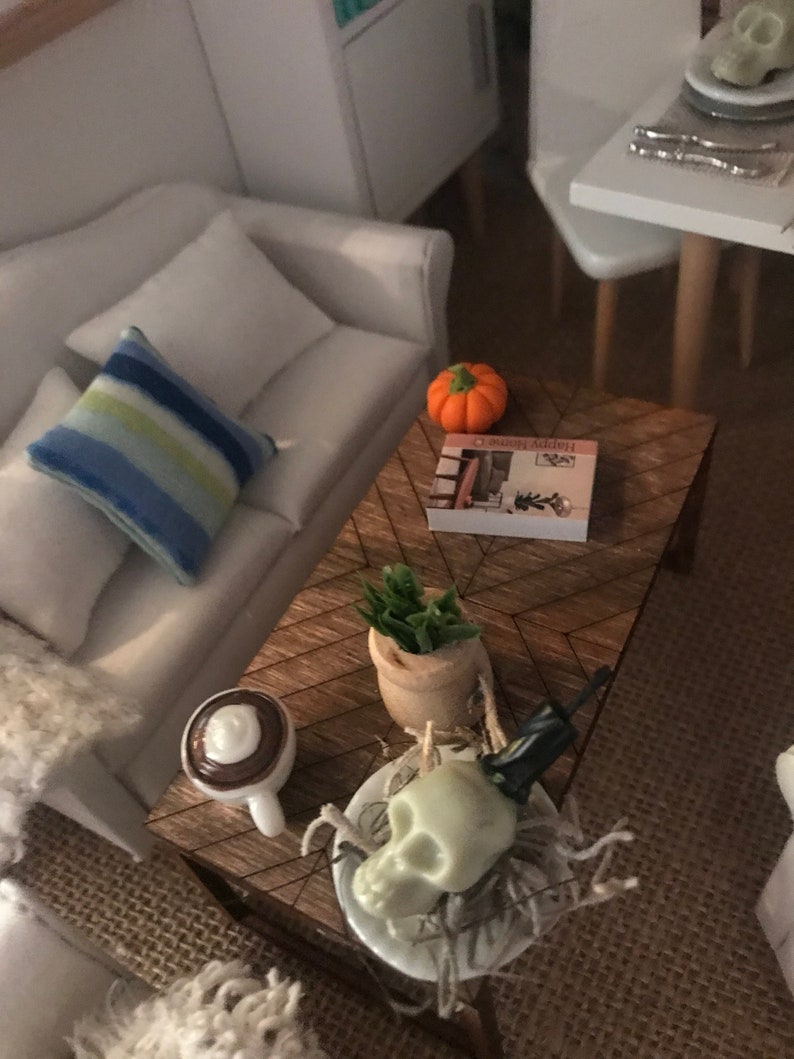 May include: Miniature living room scene with a white sofa, blue striped pillow, and a wooden coffee table. The table holds a small orange pumpkin, a book, a potted plant, a coffee cup, and a skull decoration, suggesting a Halloween theme.