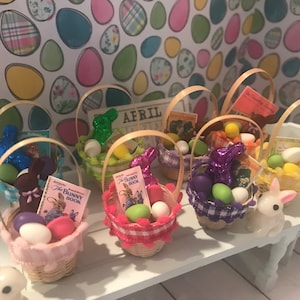 Può includere: Sei cestini pasquali in miniatura pieni di caramelle colorate, coniglietti di cioccolato e libretti. I cestini sono fatti di vimini intrecciato e hanno una varietà di nastri e decorazioni colorati. I cestini sono disposti su un tavolo bianco con una statuetta di coniglio bianco.
