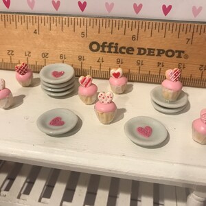May include: A collection of miniature cupcakes with pink frosting and heart-shaped toppers, alongside small grey plates with heart designs. A wooden ruler is positioned behind the items, showing a scale of approximately 12 inches in length.