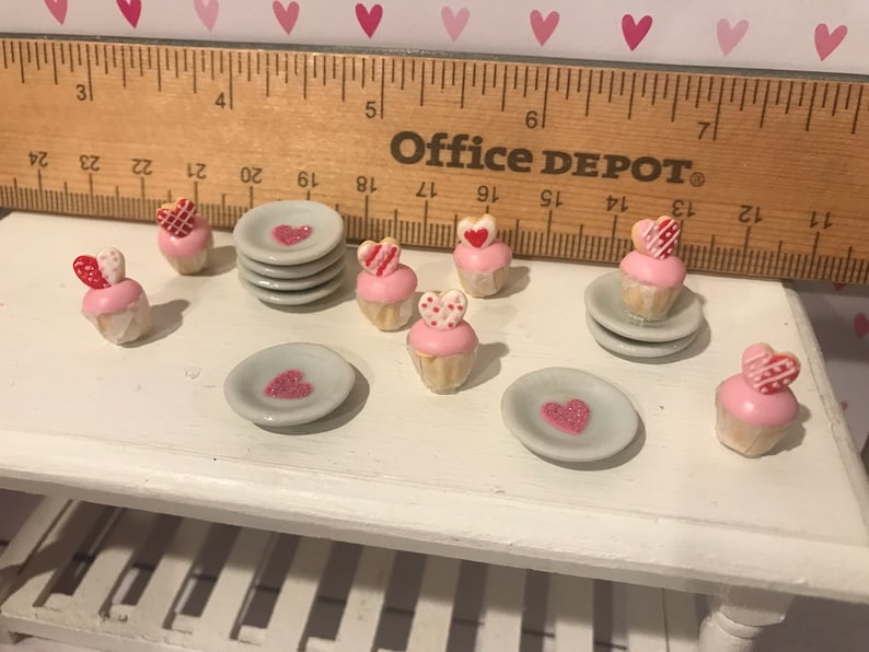 May include: A collection of miniature cupcakes with pink frosting and heart-shaped toppers, alongside small grey plates with heart designs. A wooden ruler is positioned behind the items, showing a scale of approximately 12 inches in length.