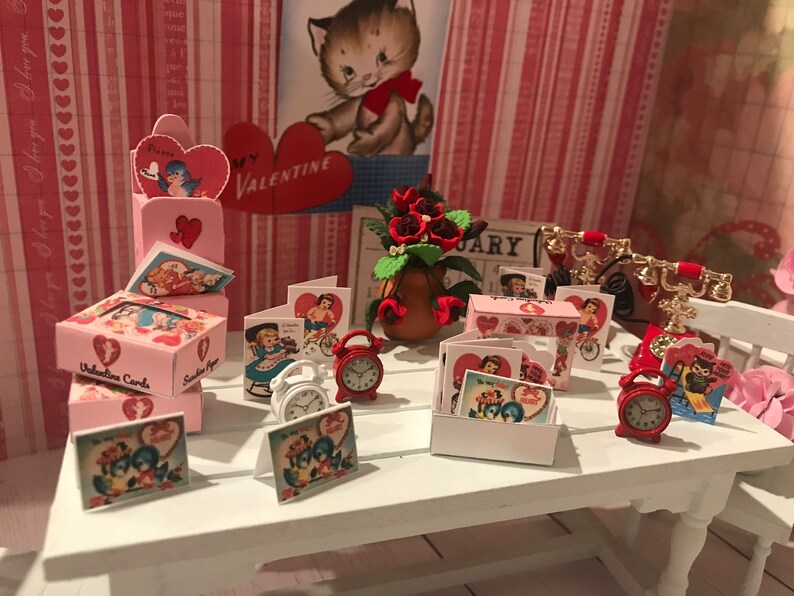 Może przedstawiać: Miniaturowa ekspozycja walentynkowa z r&oacute;żowymi pudełkami na kartki, kartkami okolicznościowymi w stylu vintage, czerwonymi budzikami i czerwonym telefonem. Obecna jest r&oacute;wnież kompozycja kwiatowa i ilustracja kota. Scena rozgrywa się na białym stole.