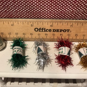 May include: A silver thimble and four bundles of Christmas tinsel in green, silver, red, and gold. Each bundle has a label with text. A wooden ruler is behind the items.
