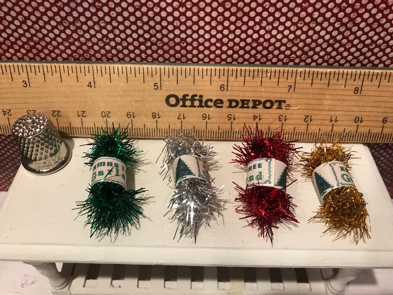May include: A silver thimble and four bundles of Christmas tinsel in green, silver, red, and gold. Each bundle has a label with text. A wooden ruler is behind the items.