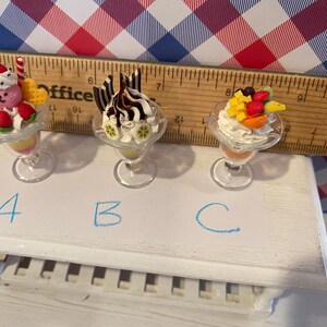 May include: Miniature dessert sundaes in clear glass dishes. One sundae has a pink bear, strawberries, and a red and white striped candy cane. Another has chocolate sauce and chocolate sticks. The third has fruit.
