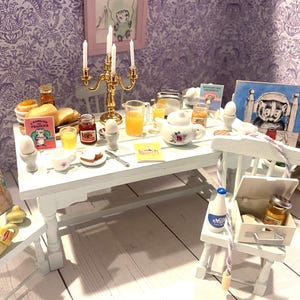 Op de afbeelding: Een miniatuur witte eettafel met een gouden kandelaar, een witte theepot met bloemmotieven, sinaasappelsap, eieren in eierdopjes, brood, jam en kleine ingelijste illustraties van dassen. Een witte stoel bevat een melkfles en een wit doosje.
