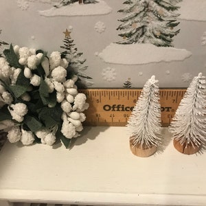 May include: Two small white flocked artificial Christmas trees with brown bases, and a white flocked floral arrangement with green leaves, all on a white shelf with a wooden ruler labeled "Office Depot" in the background.