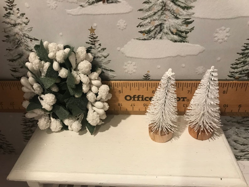 May include: Two small white flocked artificial Christmas trees with brown bases, and a white flocked floral arrangement with green leaves, all on a white shelf with a wooden ruler labeled "Office Depot" in the background.