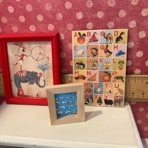 Puede incluir: Tres obras de arte en miniatura enmarcadas. Una imagen enmarcada en rojo representa una escena de circo con un elefante. Una imagen con marco de madera muestra un patr&oacute;n de osos blancos sobre un fondo azul. Tambi&eacute;n se muestra una colorida tabla del alfabeto.