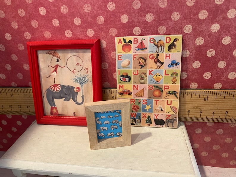 Puede incluir: Tres obras de arte en miniatura enmarcadas. Una imagen enmarcada en rojo representa una escena de circo con un elefante. Una imagen con marco de madera muestra un patr&oacute;n de osos blancos sobre un fondo azul. Tambi&eacute;n se muestra una colorida tabla del alfabeto.