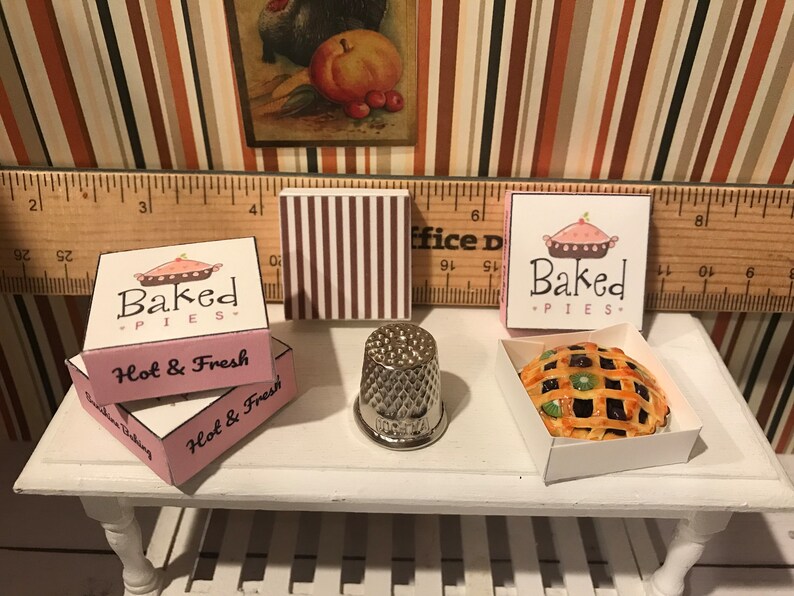 May include: Miniature baked pies with lattice crusts and fruit filling, displayed in white boxes. The boxes are labeled "Baked Pies" and "Hot & Fresh". A silver thimble and a striped box are also present.
