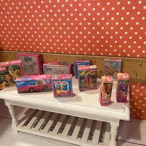 Op de afbeelding: Een verzameling miniatuur Barbie-dozen, met verschillende thema's zoals Dallas, een roze cabriolet en fietsen. De dozen staan op een kleine witte tafel tegen een rode achtergrond met stippen.