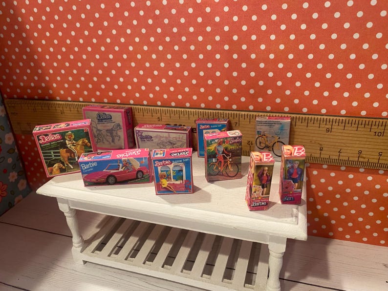 Op de afbeelding: Een verzameling miniatuur Barbie-dozen, met verschillende thema's zoals Dallas, een roze cabriolet en fietsen. De dozen staan op een kleine witte tafel tegen een rode achtergrond met stippen.