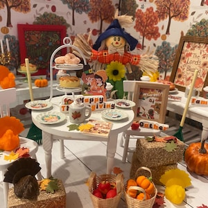 Może przedstawiać: Miniaturowa scena z okazji Święta Dziękczynienia ze strachem na wróble, nakryciami stołu i jesiennymi dekoracjami. Stół jest nakryty talerzami, wielopoziomowym stojakiem na desery i kubkiem. Dynie, bele siana i kosze owoców dodają świątecznej atmosfery.